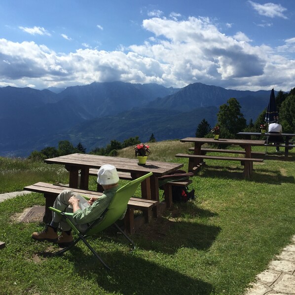 A person in a green chair reading a book at a wooden picnic table with a mountain view in the background. - Malga Zanga (Arco)