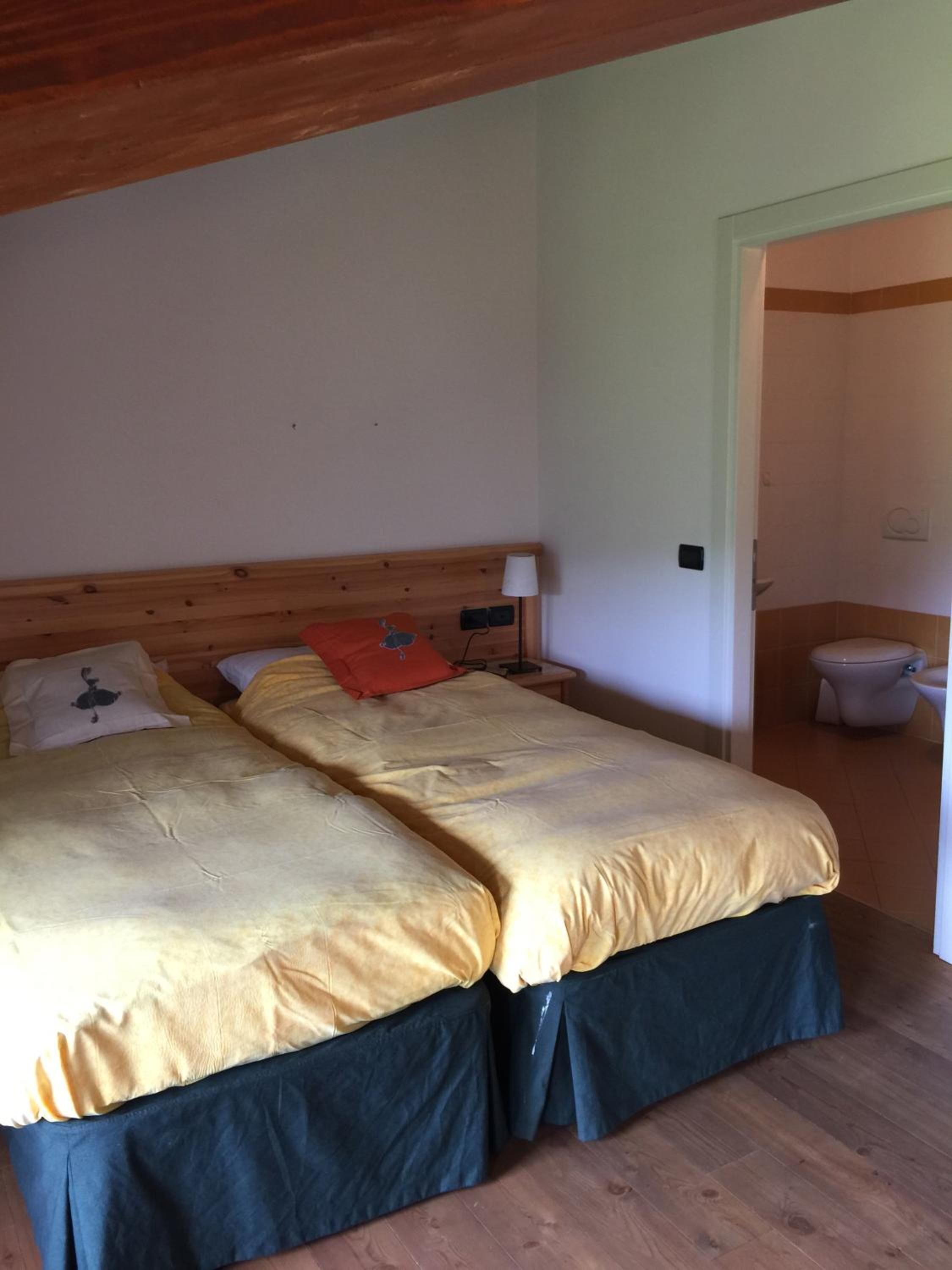 A room with two beds, a wooden ceiling, a nightstand with a lamp, and a bathroom visible through an open door.