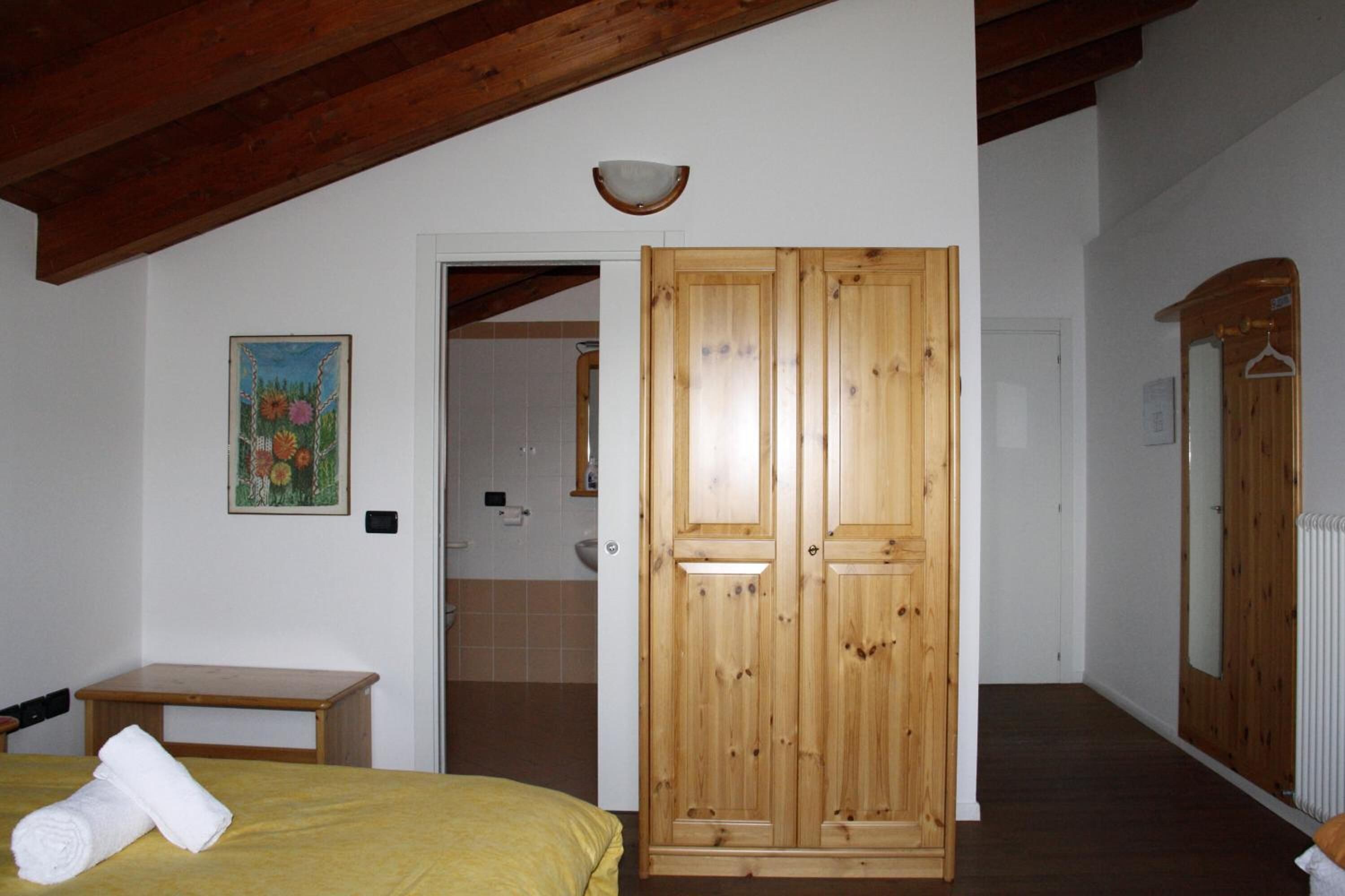 Una habitación con un armario de madera, una cama con colcha amarilla, una mesita con una toalla enrollada, un cuadro en la pared y un baño visible a través de una puerta abierta.