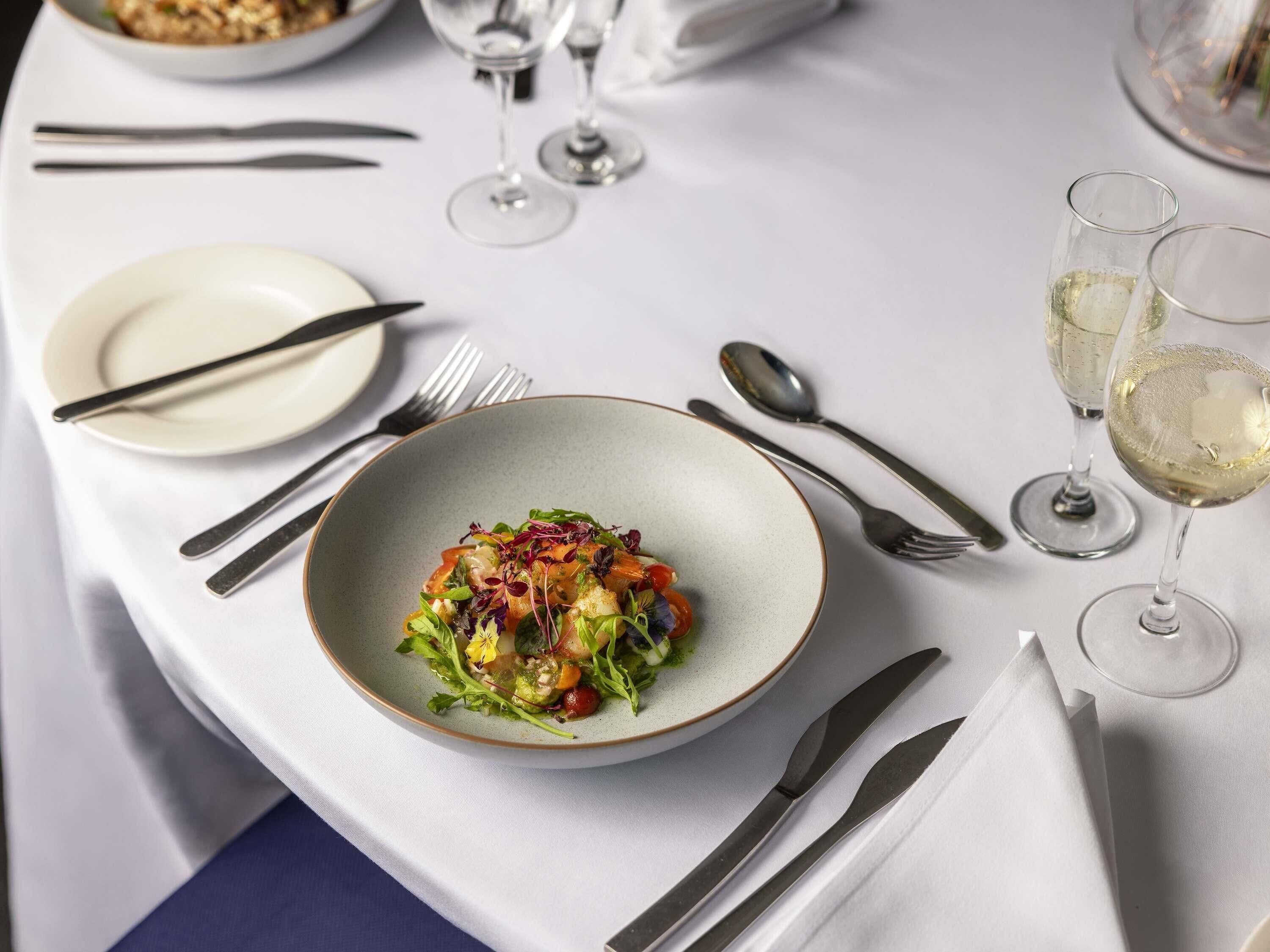 A dining table with a bowl of salad, cutlery, and glasses of white wine.