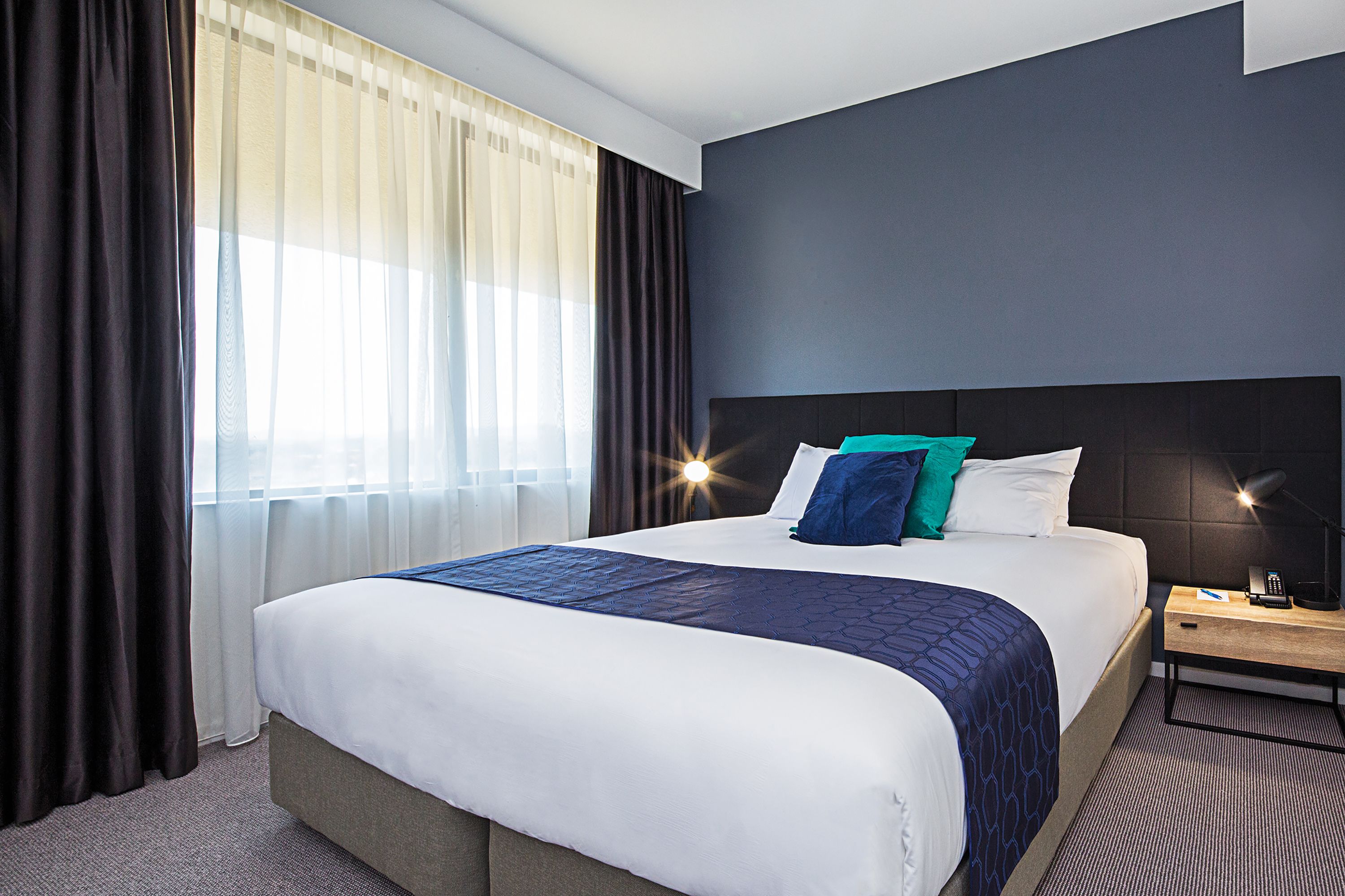 A hotel room with a large bed, a nightstand, a lamp, and a window with curtains.