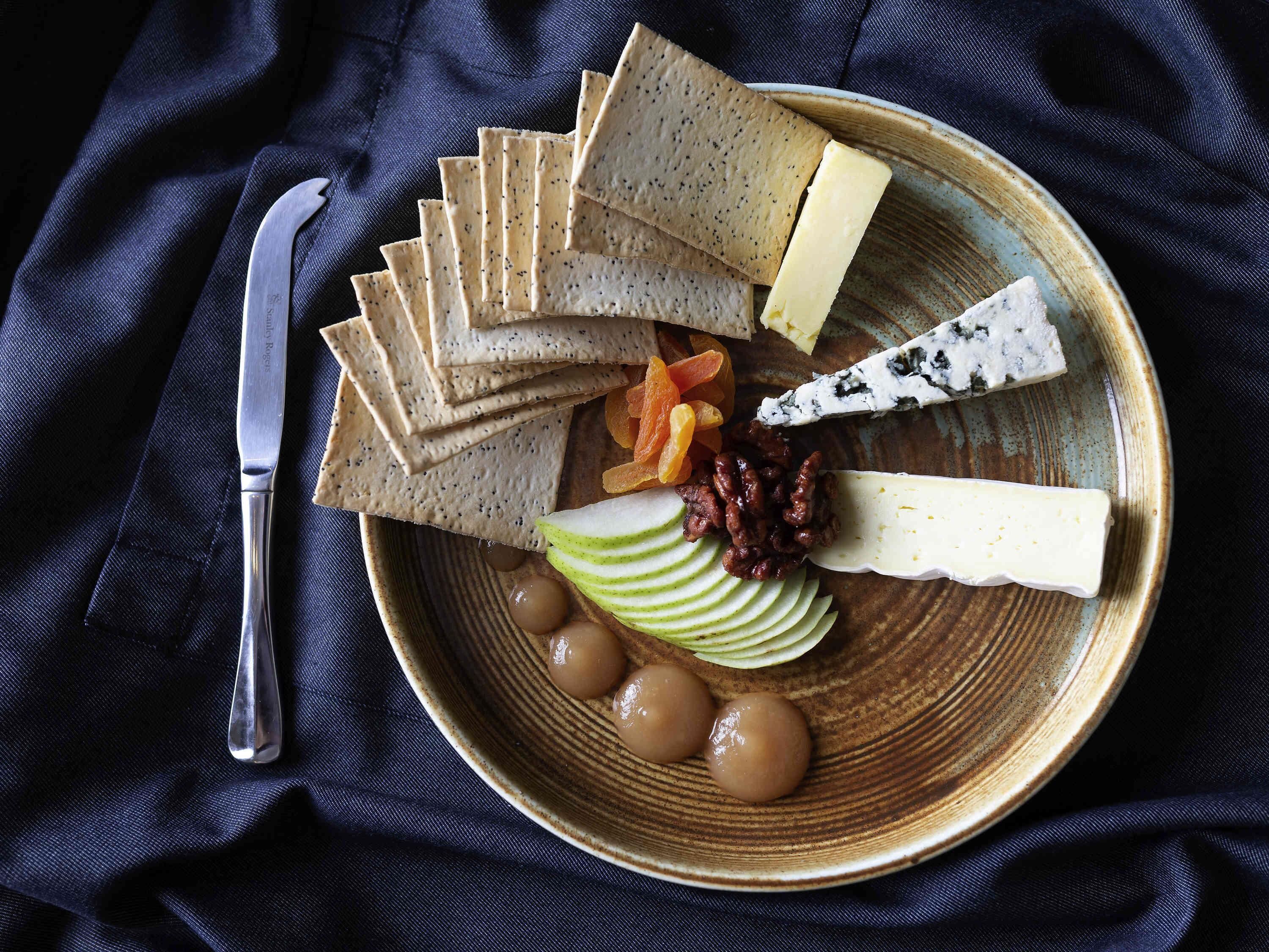 A plate of assorted cheeses, crackers, dried fruits, nuts, and sliced apples on a textured ceramic plate.