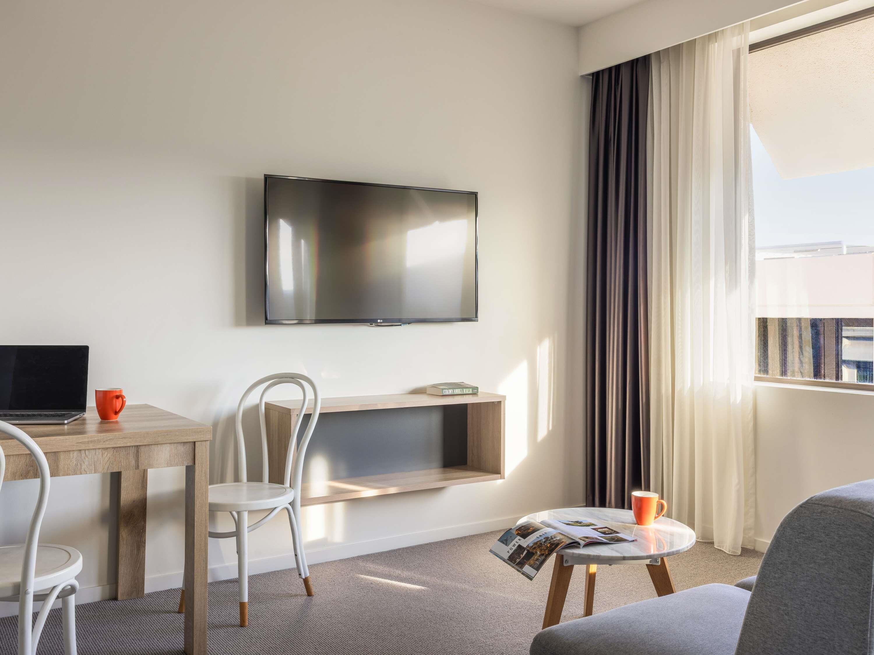 Hotellihuone, jossa on taulutelevisio, puinen työpöytä, tuoli, sohva ja ikkuna, jossa on verhot.