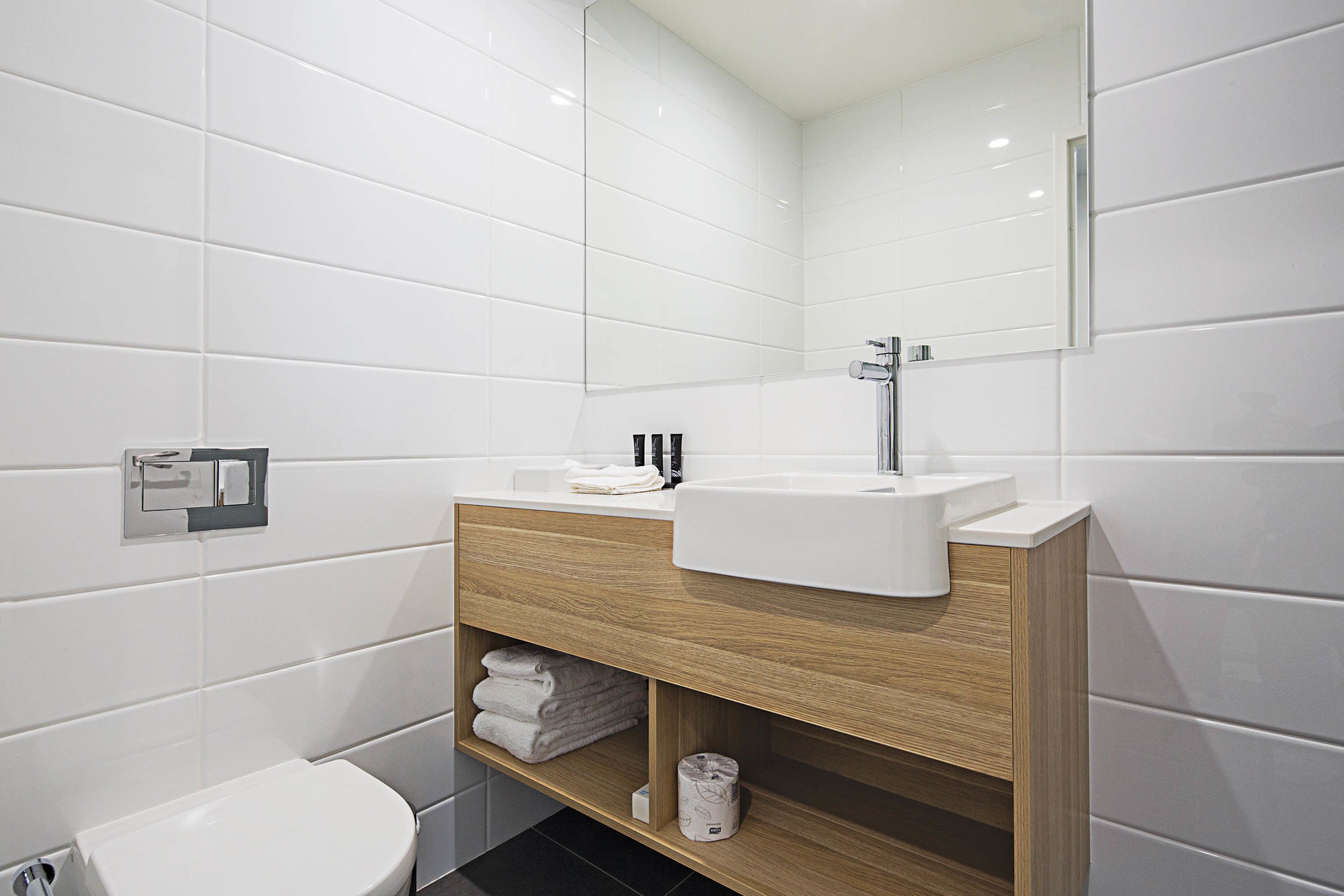 A modern bathroom with white tiles, a wooden vanity, a white sink, and a toilet.