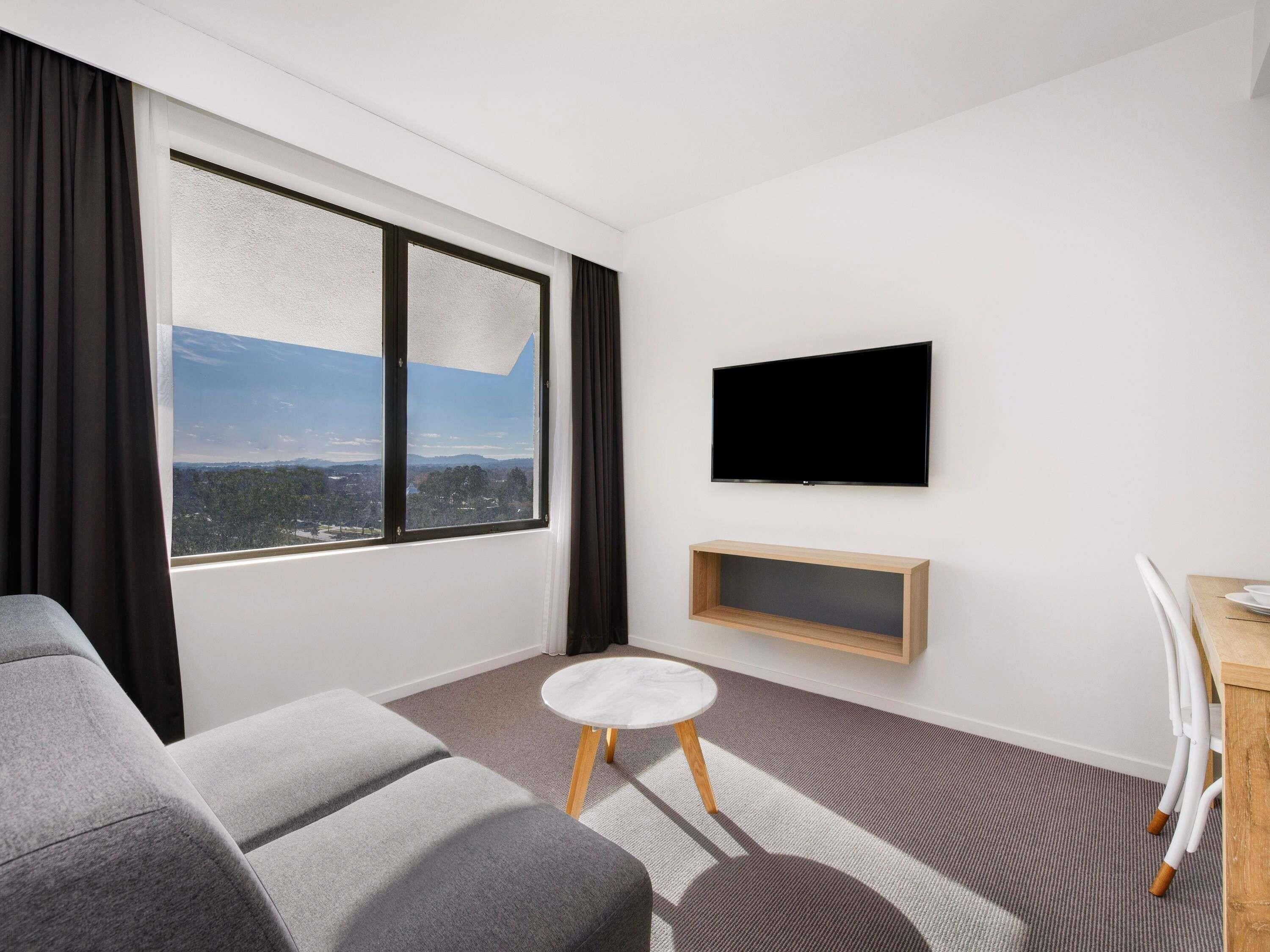 Una habitación moderna con un ventanal grande, un televisor de pantalla plana, un sofá, una mesita redonda y un mueble bajo de madera para el televisor.