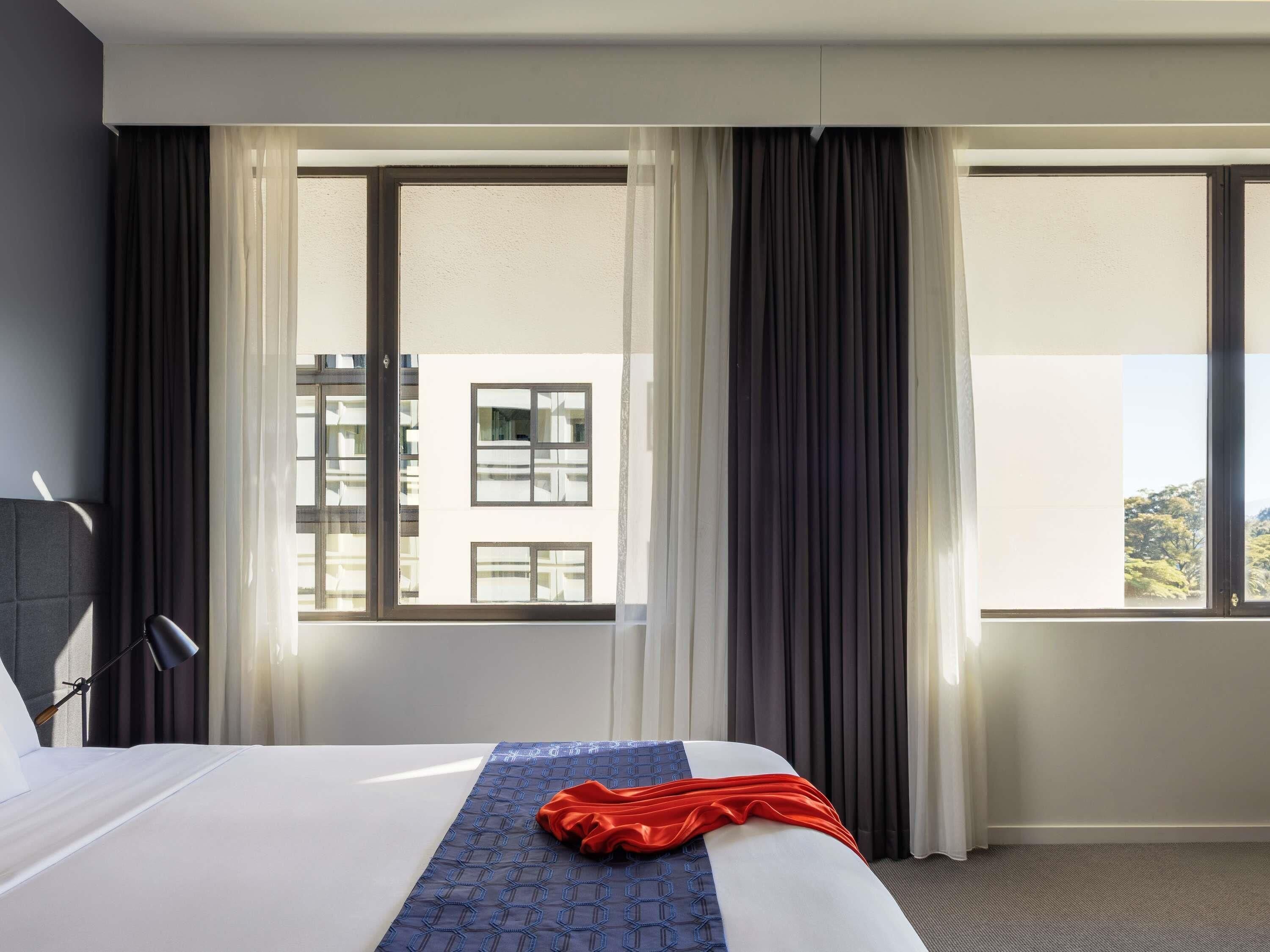 A hotel room with a bed, a window with curtains, and a view of buildings outside.