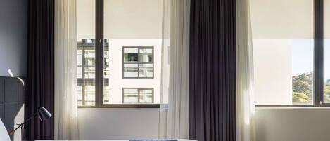 A hotel room with a bed, a window with curtains, and a view of buildings outside.
