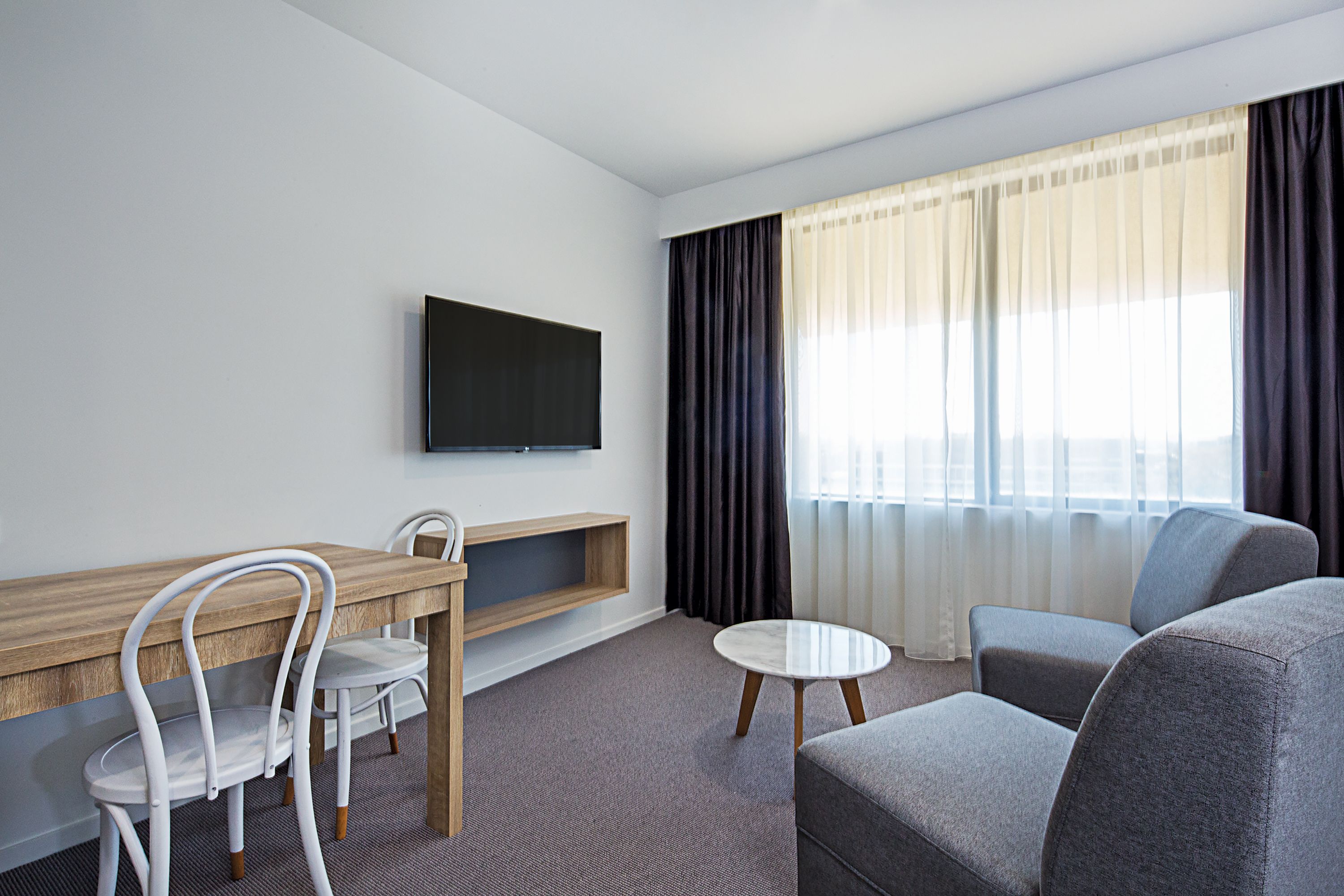 Habitación de hotel moderna con un escritorio de madera, dos sillas blancas, un sofá gris, una mesa redonda blanca y un televisor de pantalla plana montado en la pared.
