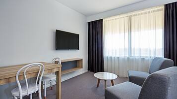 Habitación de hotel moderna con un escritorio de madera, dos sillas blancas, un sofá gris, una mesa redonda blanca y un televisor de pantalla plana montado en la pared.