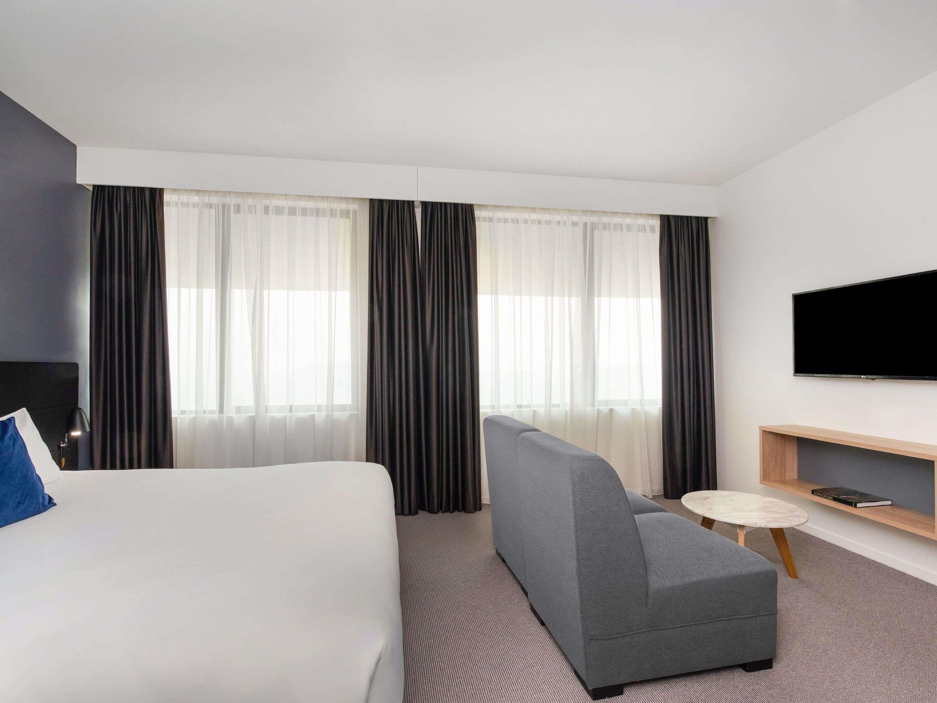 A hotel room with a bed, a grey sofa, a small round table, a TV mounted on the wall, and a bookshelf.