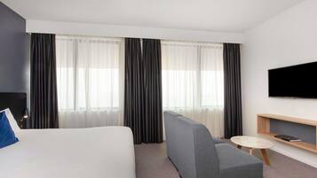 A hotel room with a bed, a grey sofa, a small round table, a TV mounted on the wall, and a bookshelf.