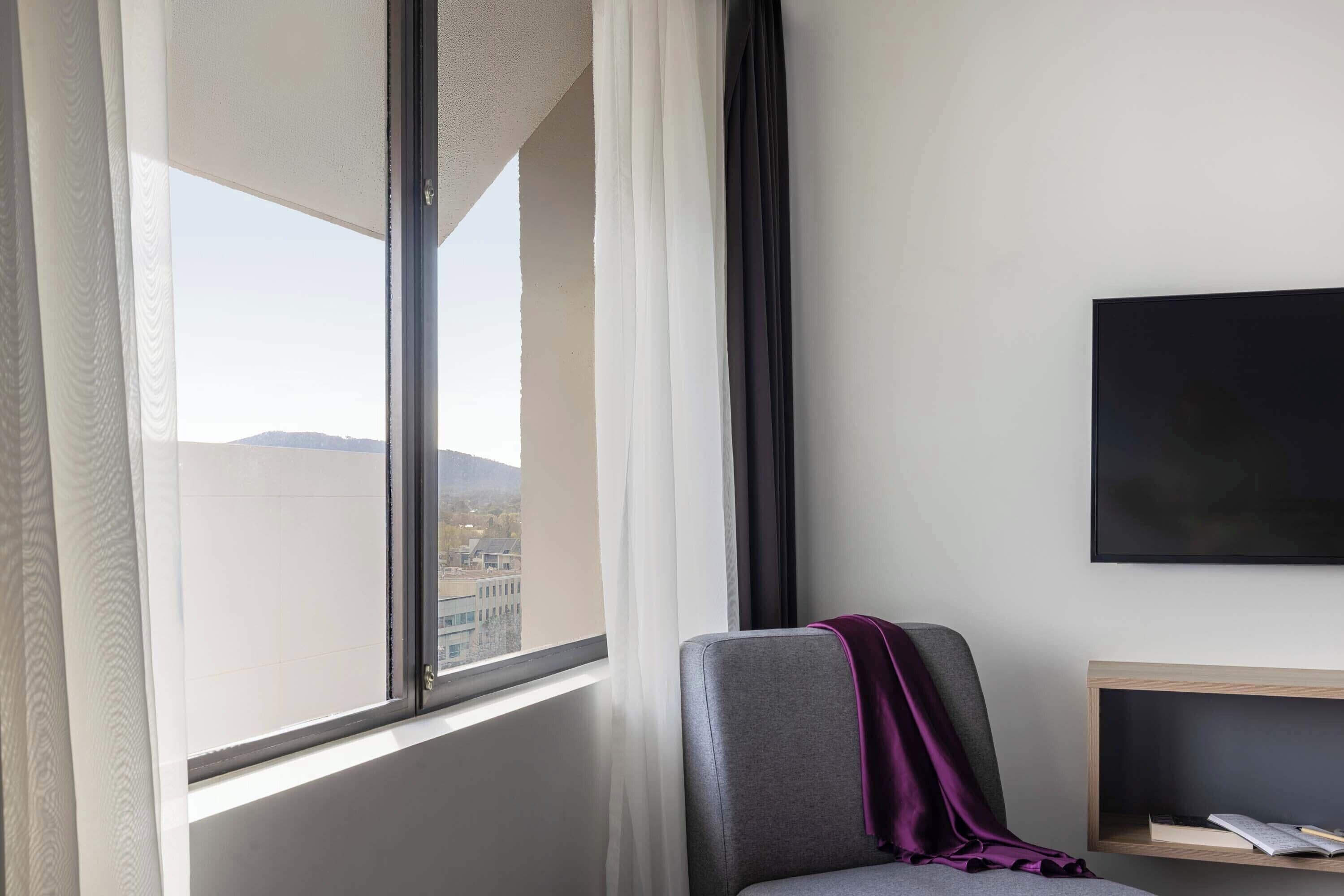Een hotelkamer met een groot raam, een grijze stoel met een paarse plaid, een flatscreen televisie en een houten bijzettafeltje.
