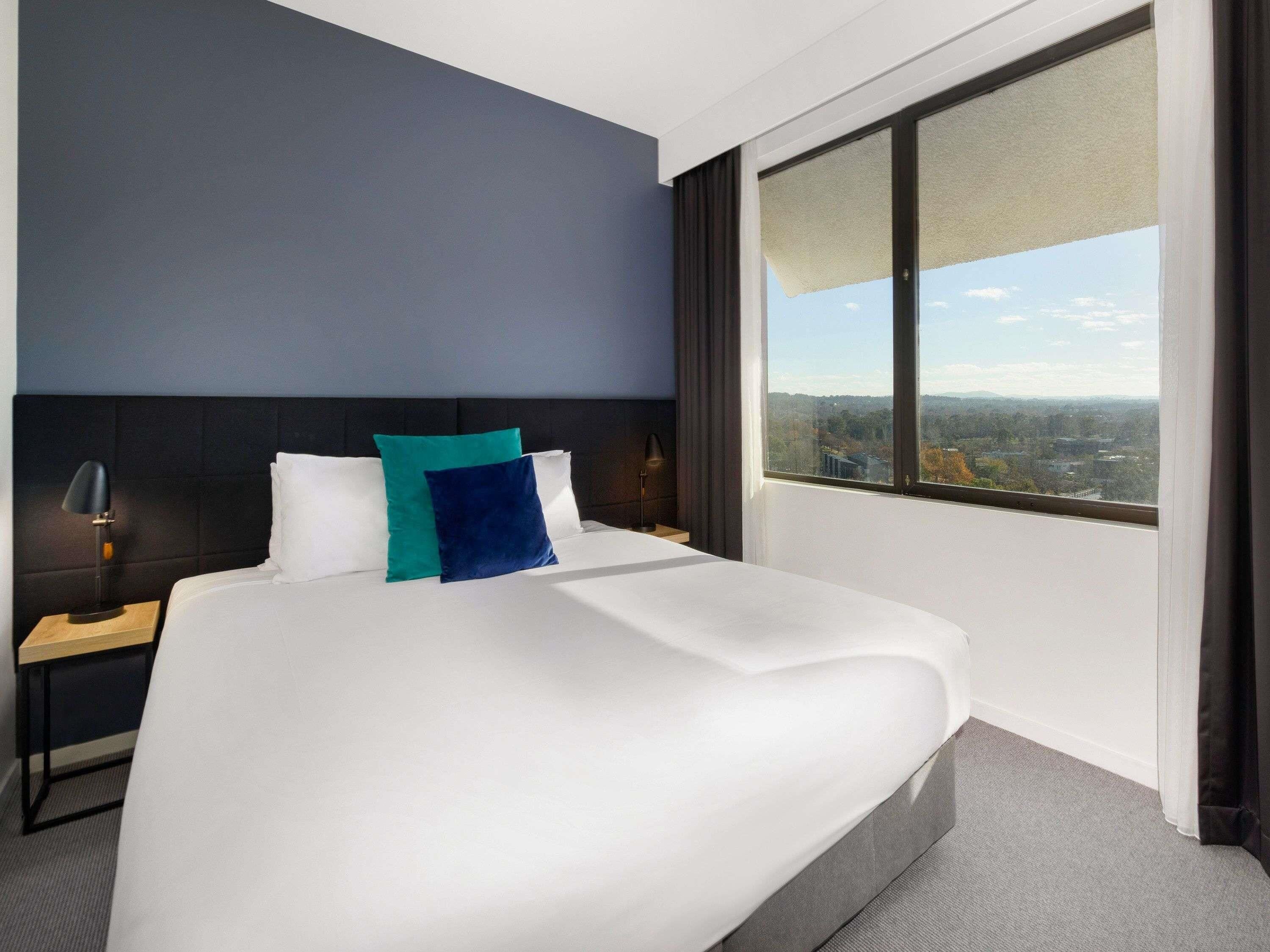 A hotel room with a large bed, a nightstand, a lamp, and a window with a city view.