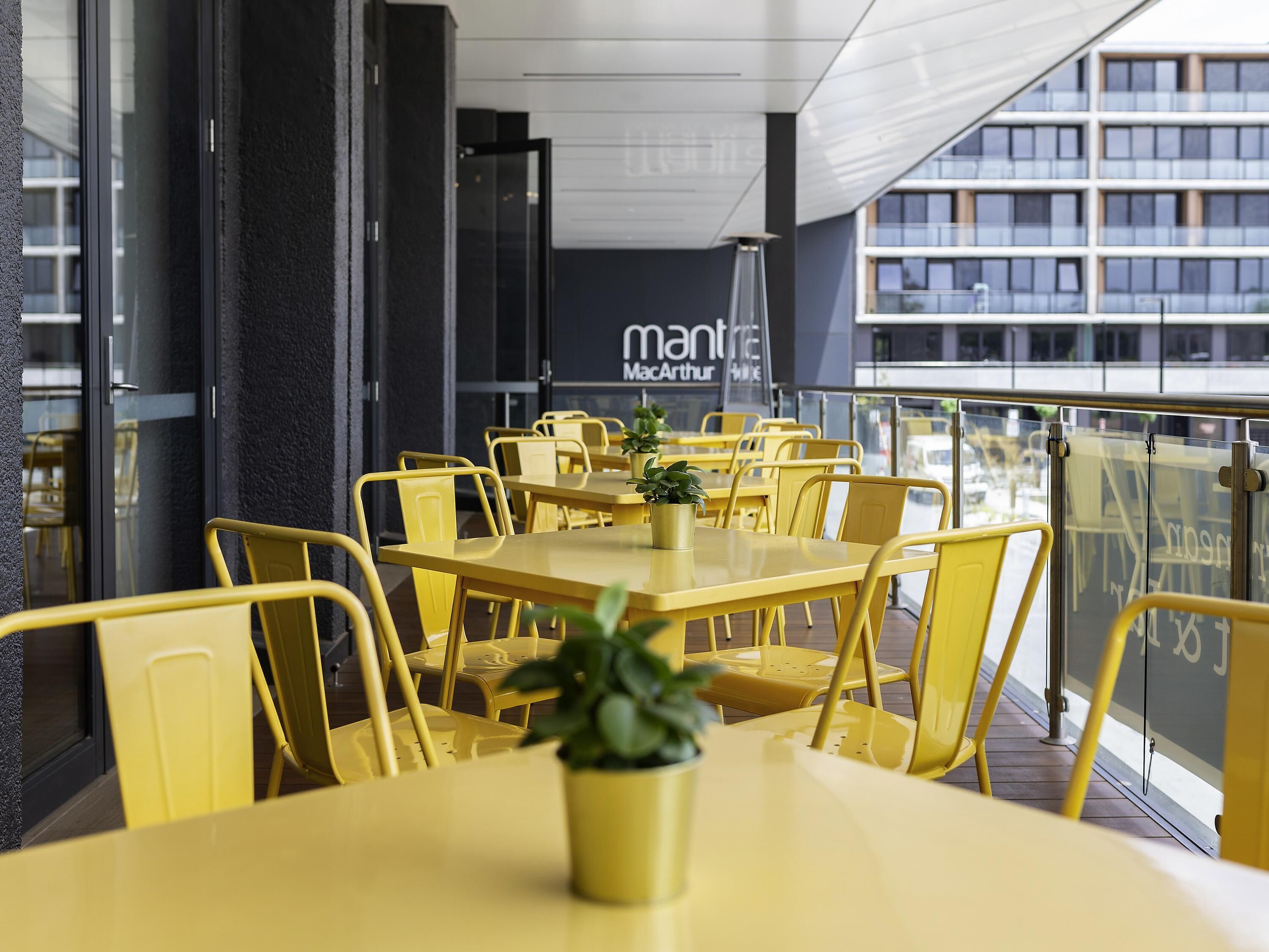 A restaurant with yellow tables and chairs, potted plants, and a view of a building with 'mantra' signage.