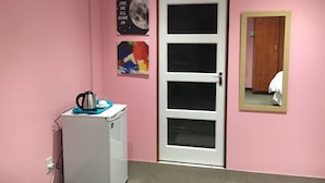 A room with a pink wall, a white door, a small white refrigerator, and a mirror on the wall. - Tassili Lodge (Kempton Park)
