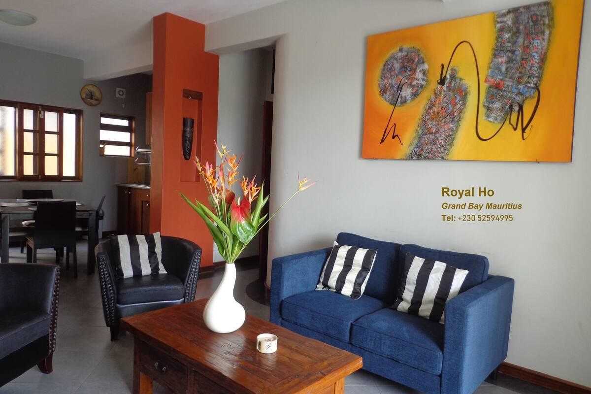 Un salon moderne avec un canapé bleu, une table basse en bois et un vase rempli de fleurs. Un tableau est accroché au mur, ainsi qu’une enseigne indiquant le nom de l’hôtel et ses coordonnées.