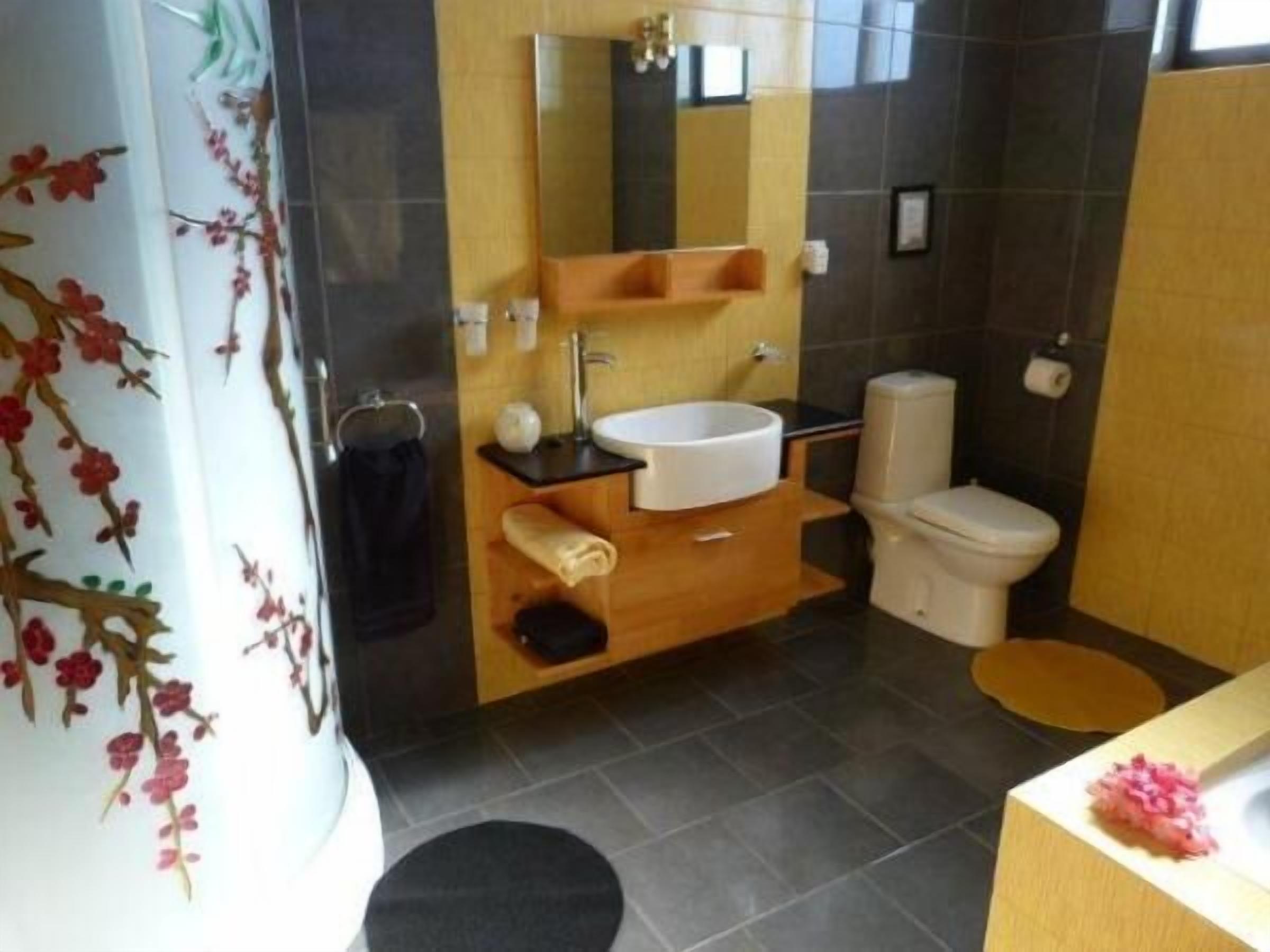 Un baño con un lavamanos blanco, un inodoro y un espejo sobre el lavamanos. Hay un perchero y una pequeña estantería con artículos de aseo.