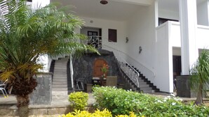 A building with a stone staircase leading to a wooden door, surrounded by greenery and a white exterior. - Royal Ho (Grand-Baie)