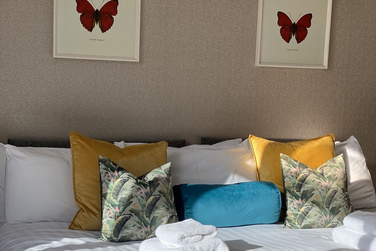 A neatly made bed with white linens, a stack of white towels, and a variety of colorful pillows.