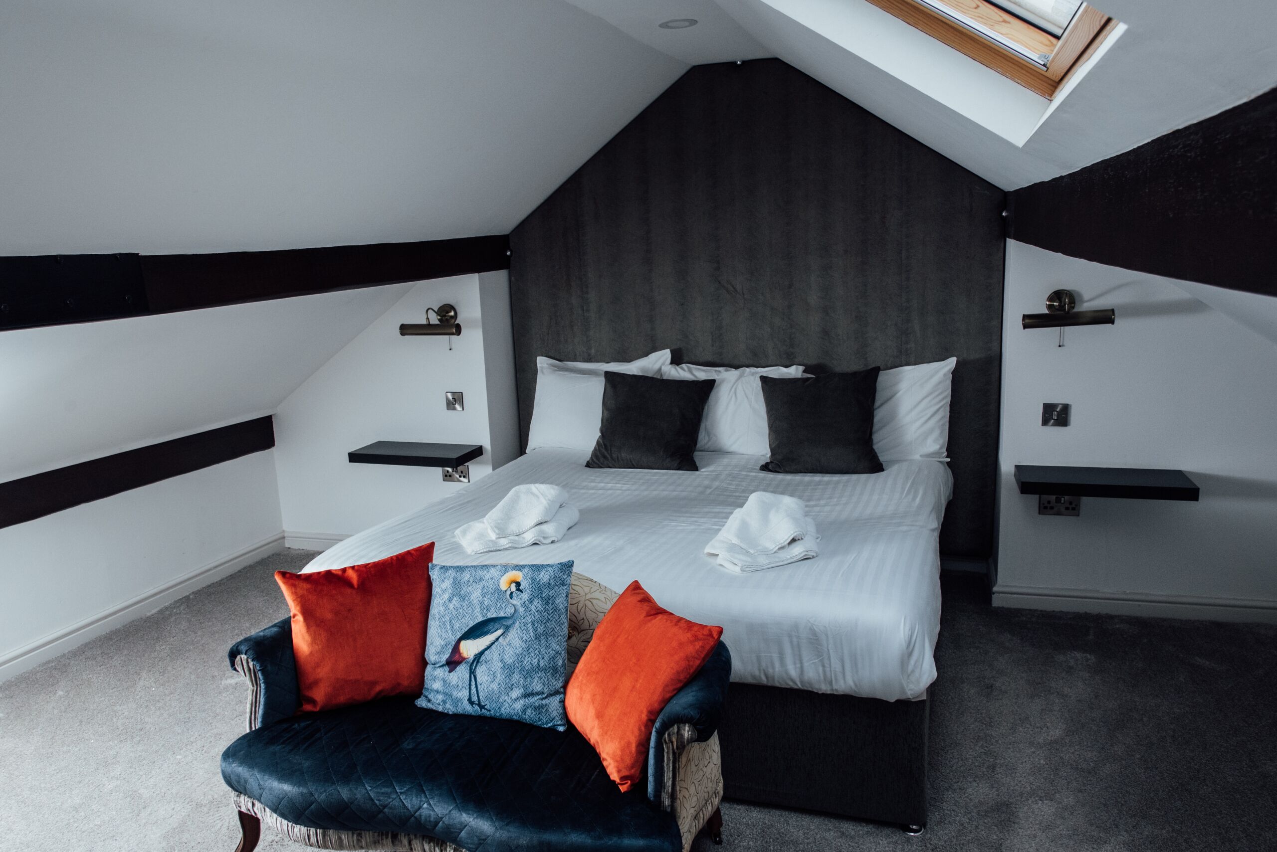 A modern bedroom with a sloped ceiling, a large bed with white linens, two bedside tables, and a chair with colorful cushions.