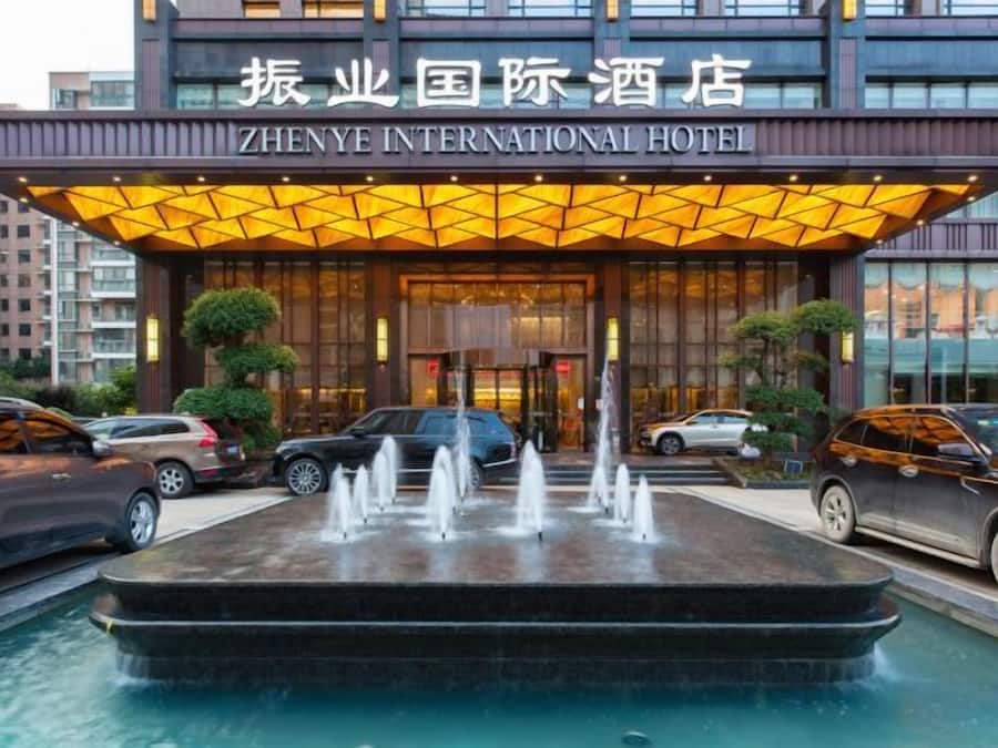 A hotel entrance with a fountain, cars parked in front, and a sign reading 'ZHENYE INTERNATIONAL HOTEL'.