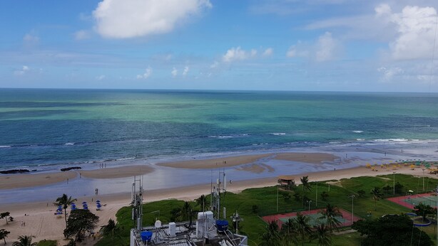 Une plage avec une eau d’un bleu limpide, un rivage sablonneux et quelques personnes. On y trouve des palmiers et un bâtiment doté d’antennes.