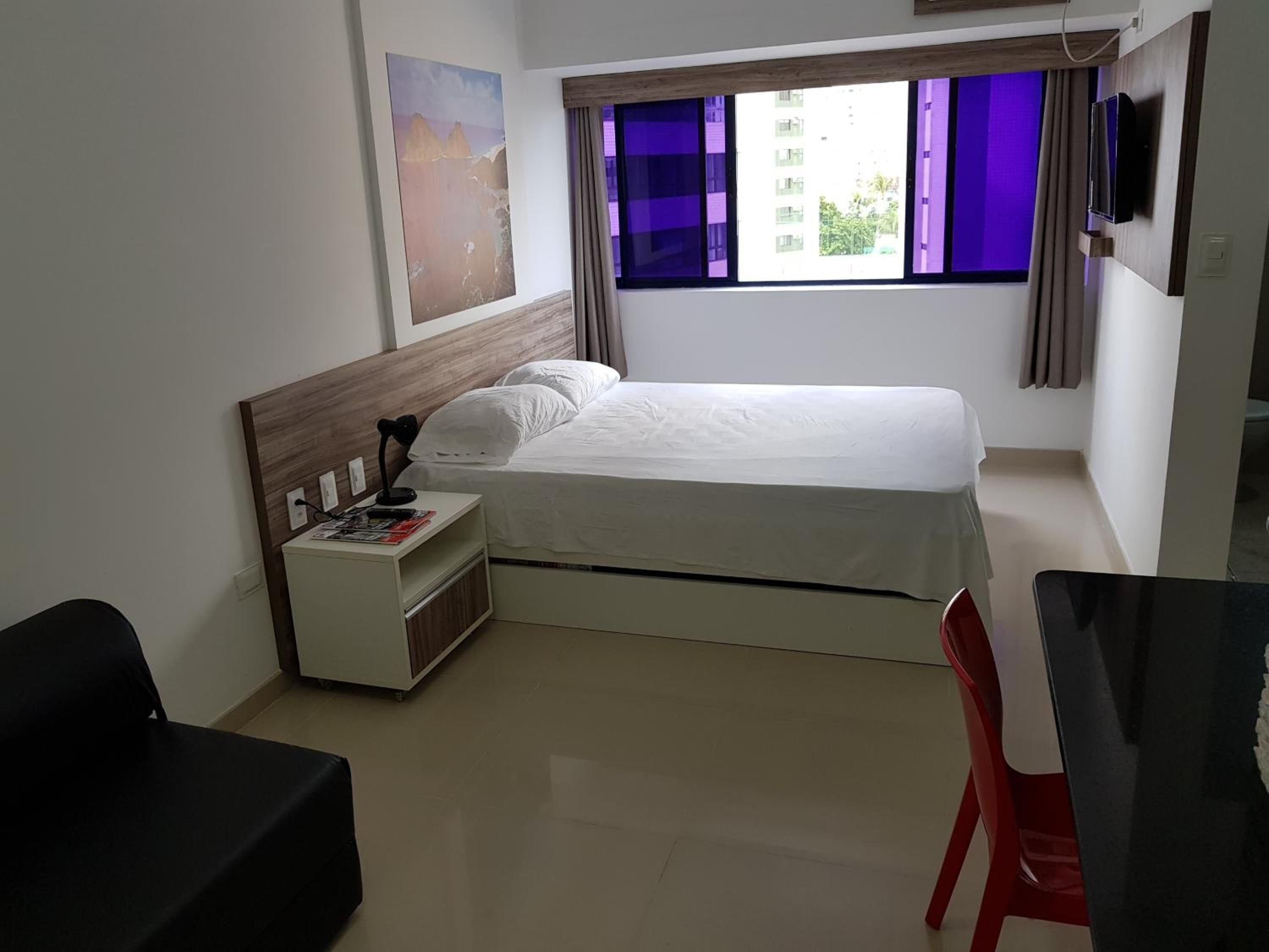 A bedroom with a bed, a nightstand, a TV, a window with purple curtains, and a painting on the wall.