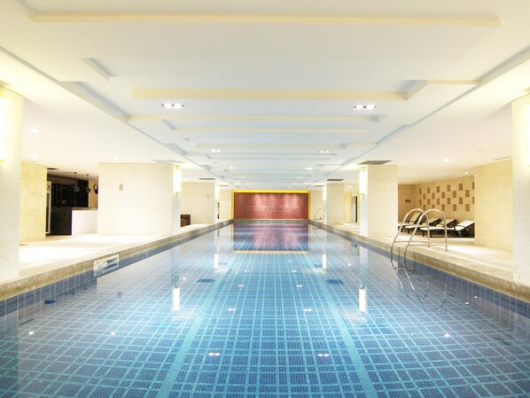 A large indoor swimming pool with a tiled floor, a reception area with a red accent wall, and seating by the pool. - Tang Dynasty West Market Hotel (Xi'an)