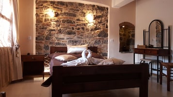 Un dormitorio con pared de piedra, una cama con muchas almohadas, una mesita de noche de madera, un aparador, un espejo y una silla.