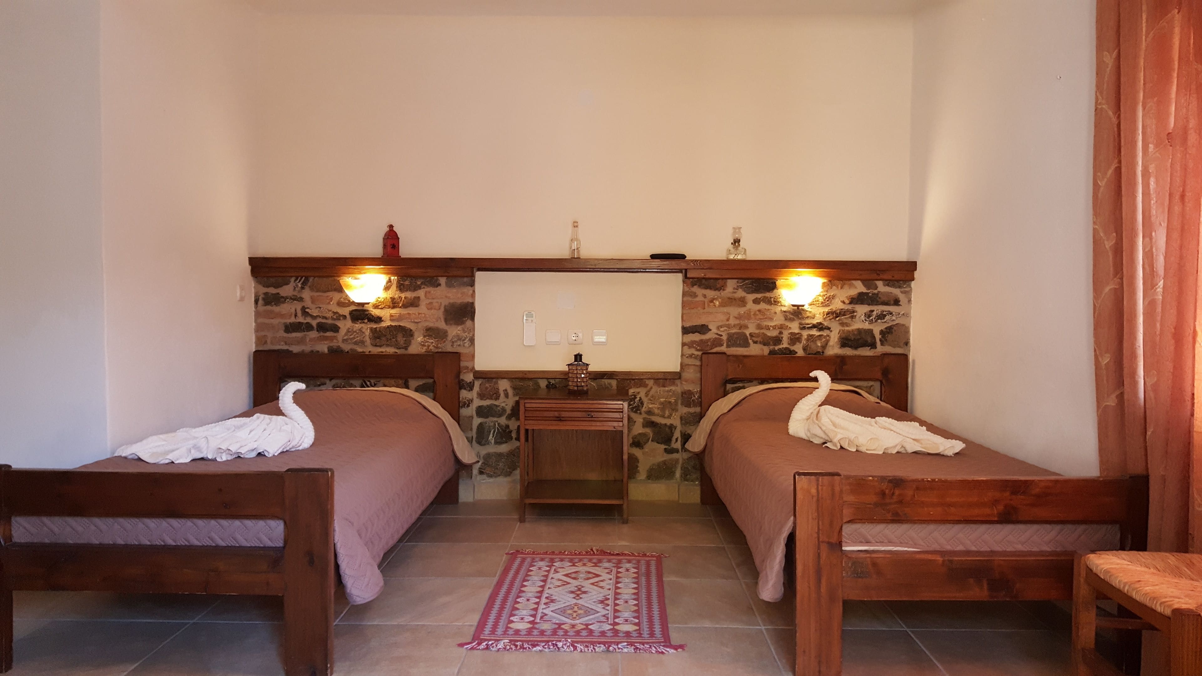 Una habitación con dos camas de madera, una pared de piedra, una mesita de noche de madera, una alfombra roja con motivos y dos luces fijadas a la pared.