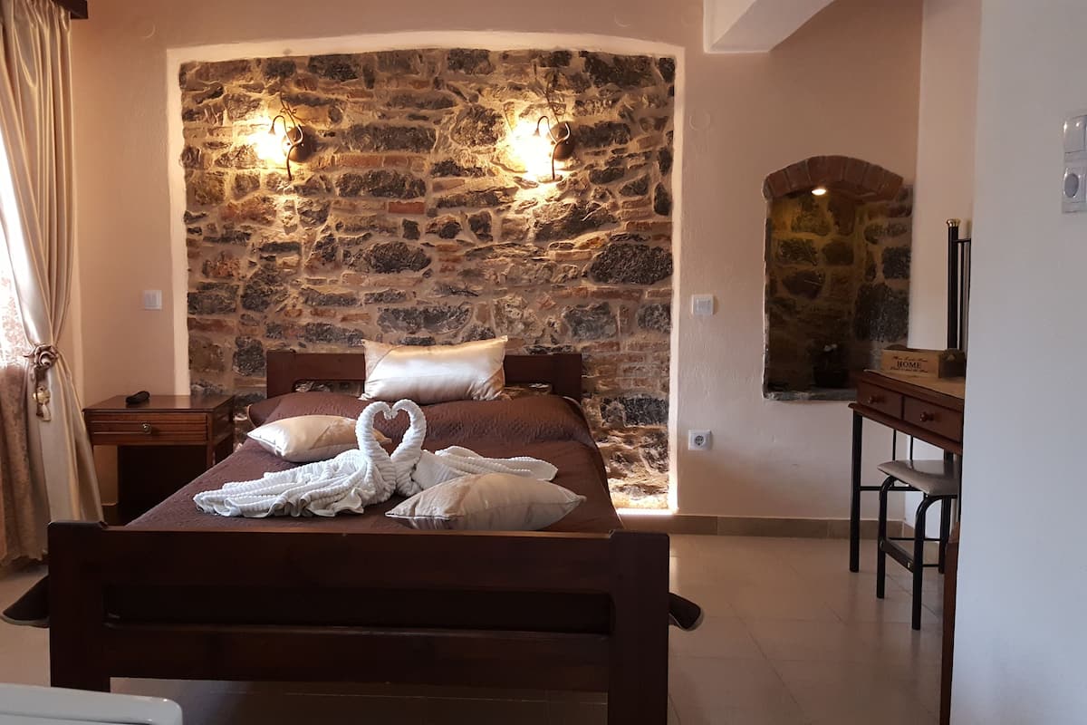 A bedroom with a stone wall, a wooden bed, a nightstand, a bedside table, a mirror, and a window with curtains.