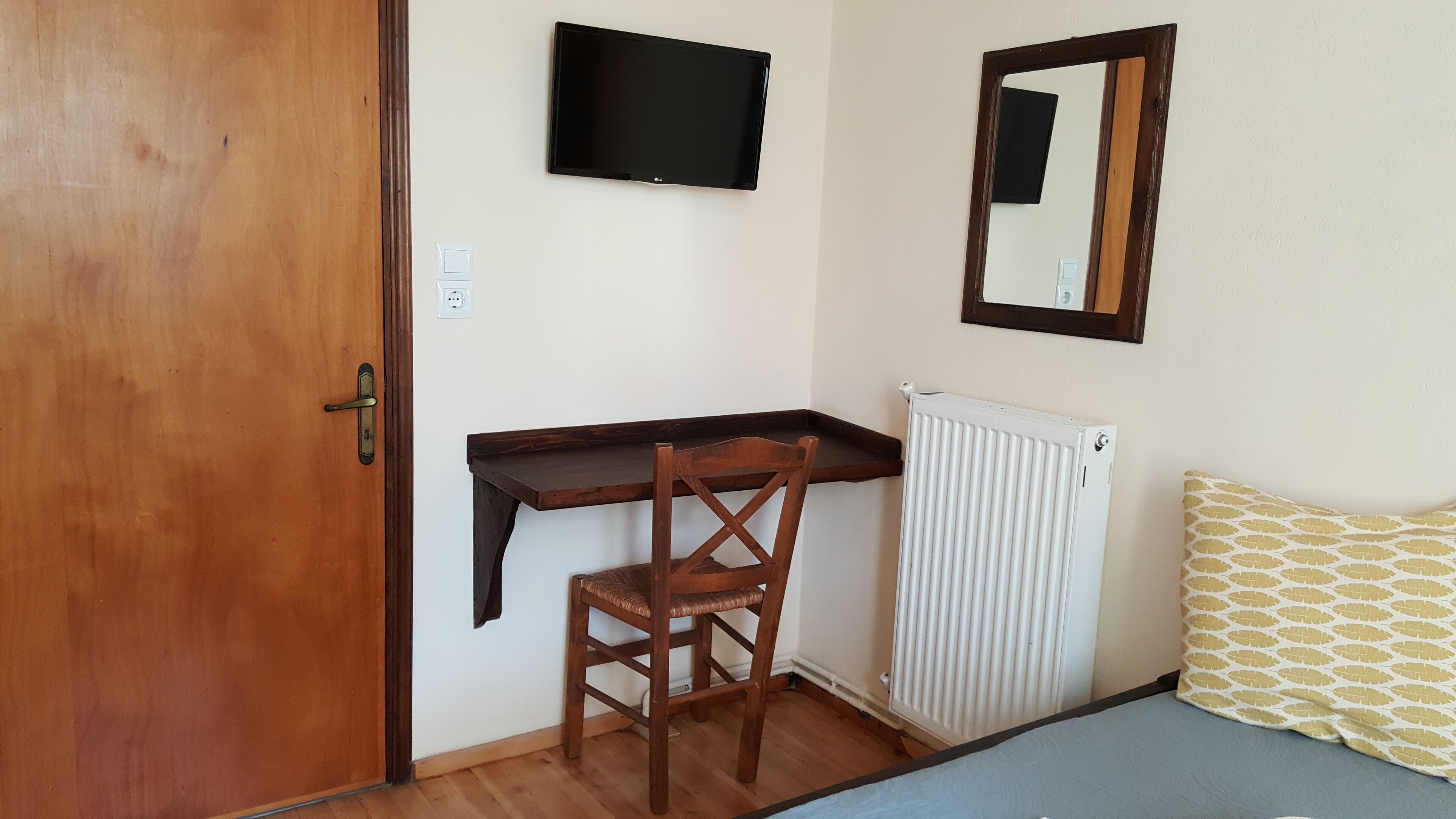 Una habitación con puerta de madera, un escritorio con silla, un televisor montado en la pared, un radiador y un cojín estampado en la cama.