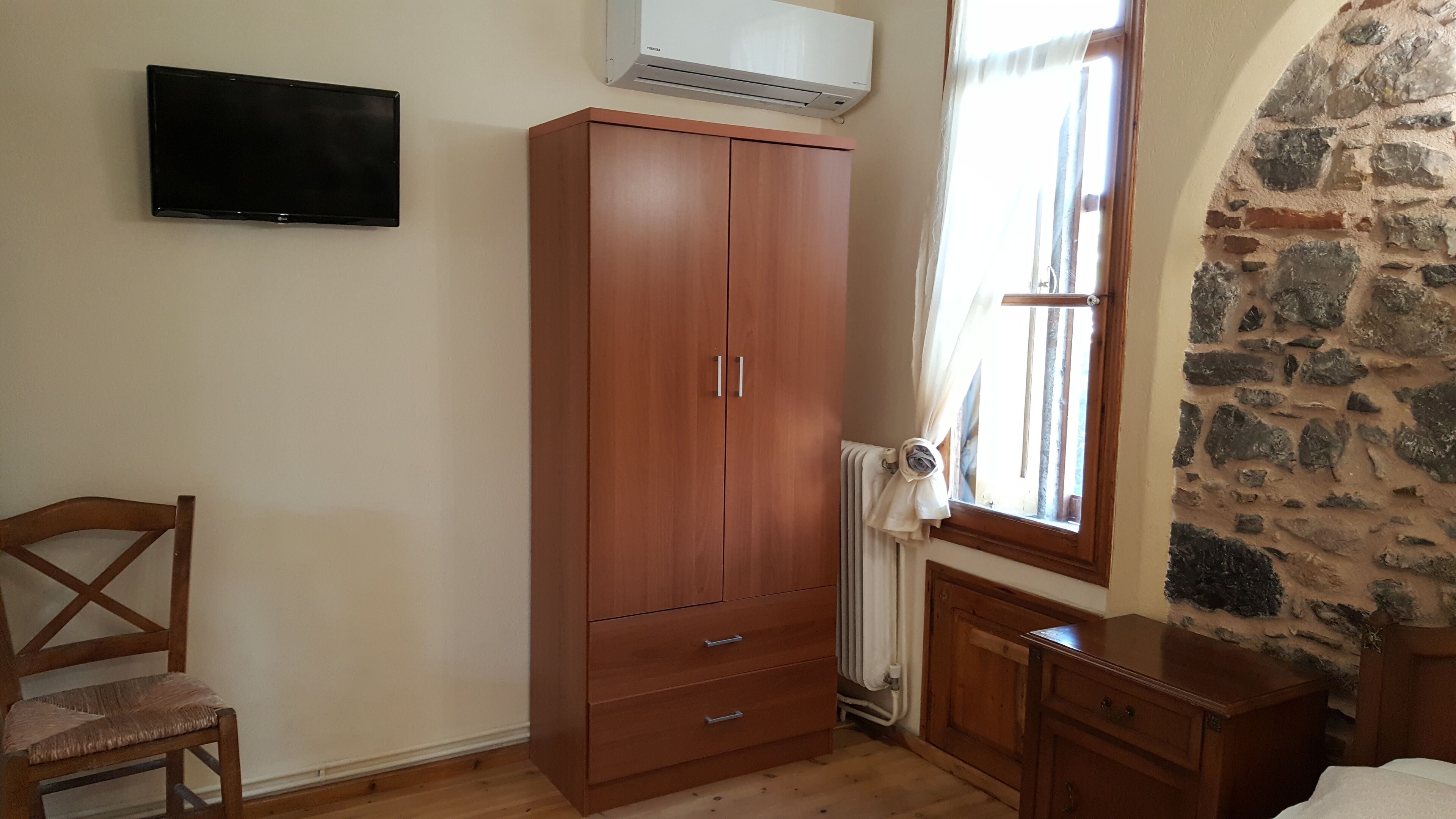 Ein Zimmer mit einem Holzschrank, einem an der Wand befestigten Fernseher, einem Fenster mit Vorhängen und einem Heizkörper.