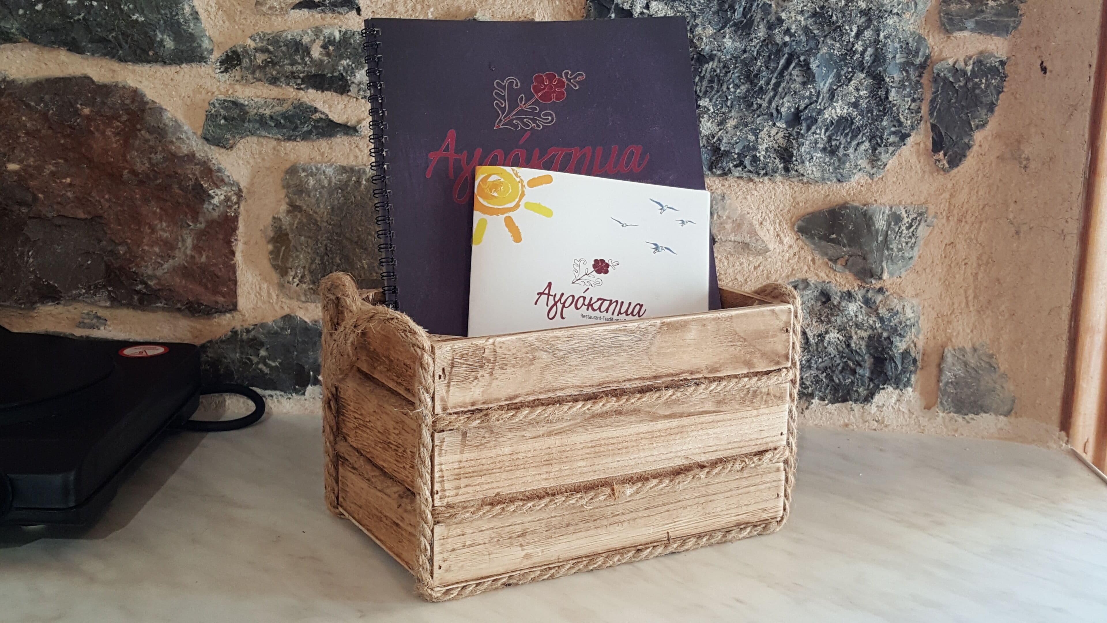Una caja de madera rústica con un libro y una tarjeta con el nombre 'Ayo' sobre un fondo de pared de piedra.