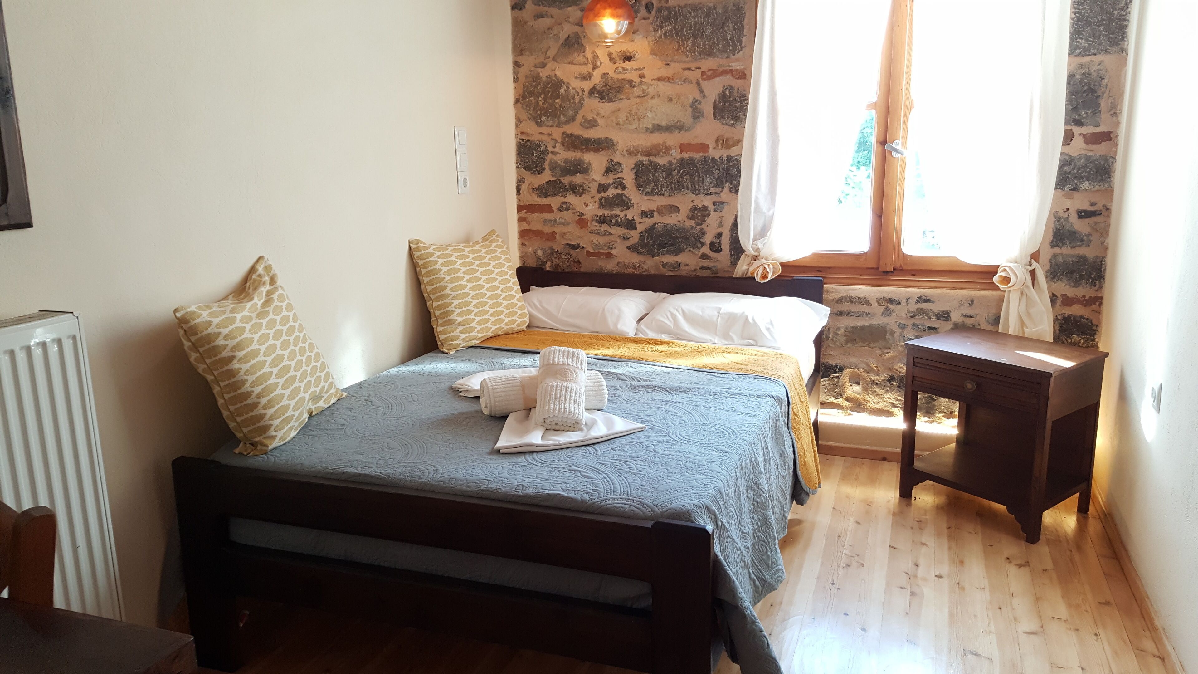 A bedroom with a bed, a wooden nightstand, a radiator, and a stone wall.