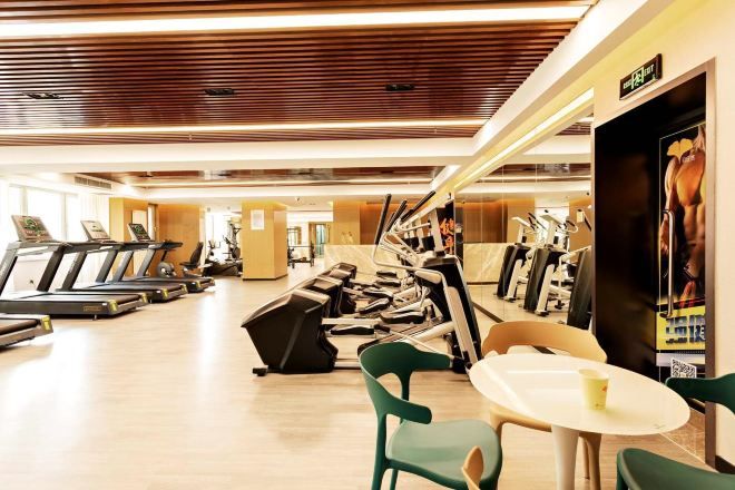 Un gimnasio con cintas de correr, bicicletas estáticas y máquinas de pesas. Hay sillas verdes y una pequeña mesa con una taza.