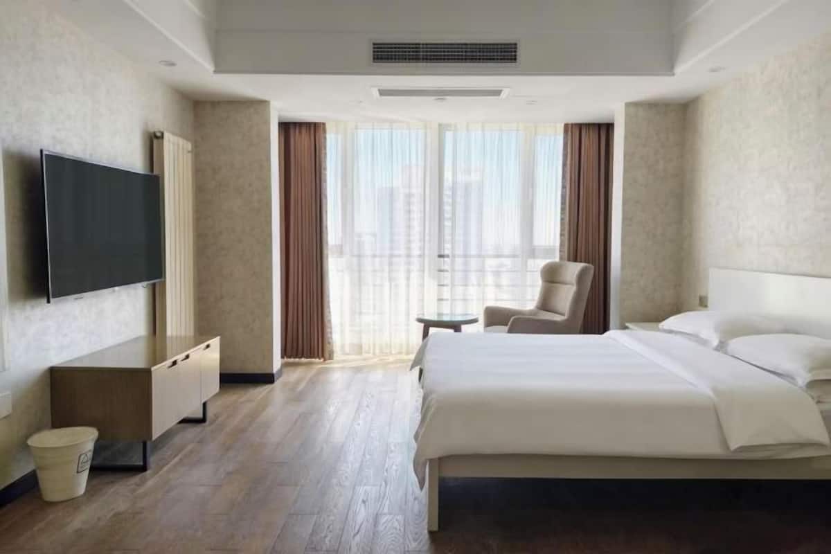 A hotel room with a bed, a TV, a chair, a small table, and a view of the city through large windows.