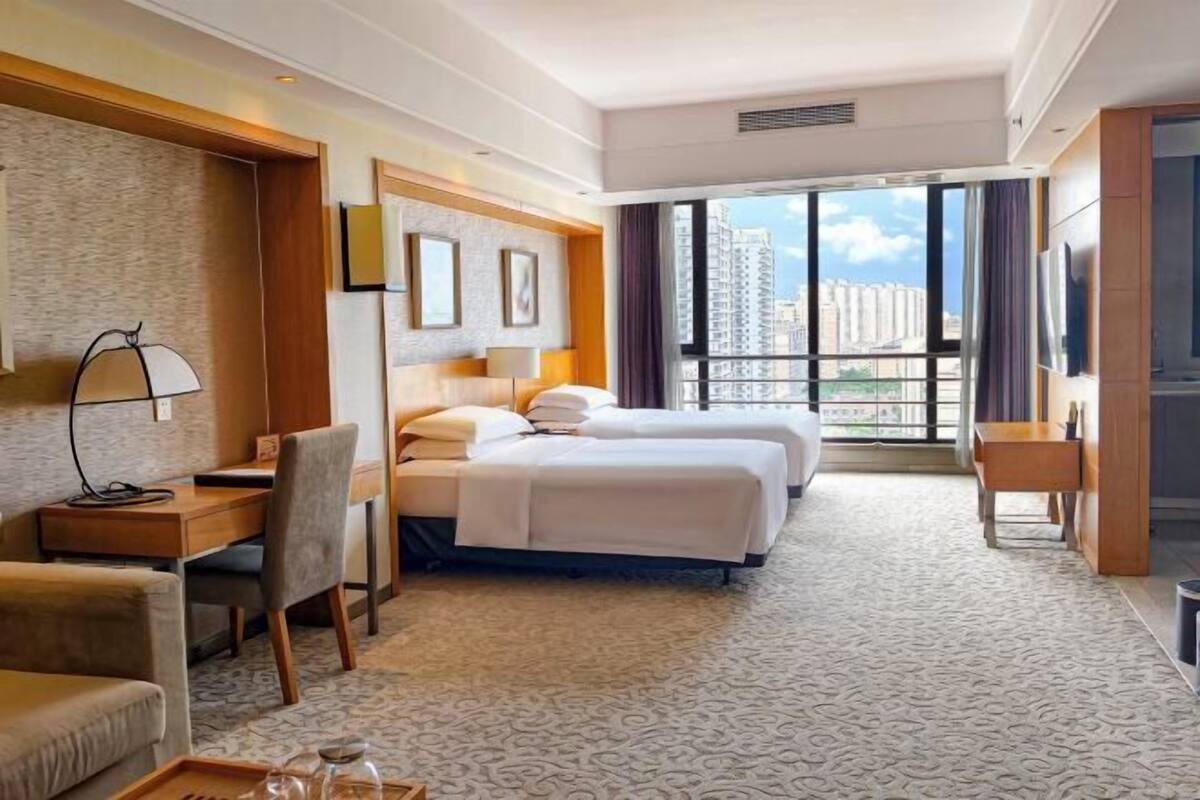 A hotel room with two beds, a desk, a chair, a TV, and a large window with a city view.