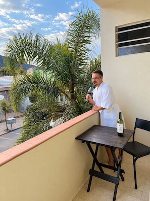 Um homem numa varanda com uma mesa, uma cadeira, uma garrafa de vinho e uma palmeira ao fundo.