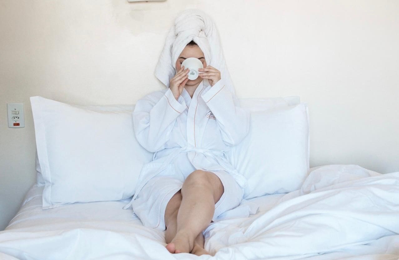 A person in a white bathrobe sitting on a bed with white bedding, a white pillow, and a white blanket.