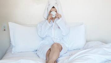 A person in a white bathrobe sitting on a bed with white bedding, a white pillow, and a white blanket.