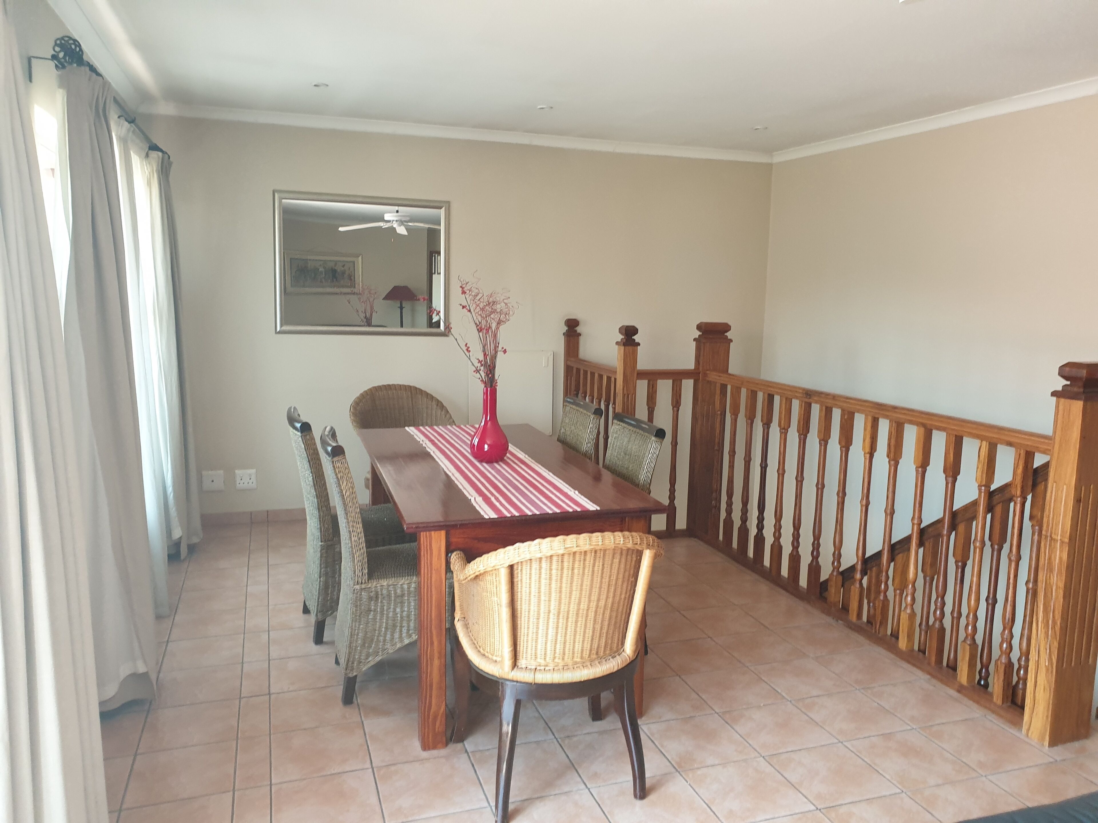 A dining area with a wooden table, wicker chairs, a red vase, and a wooden railing.