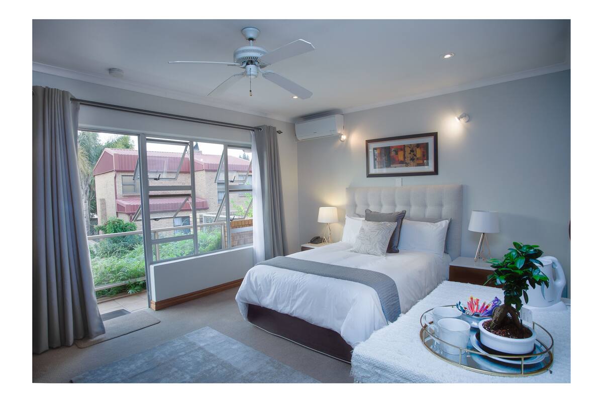 Una habitación de hotel con cama, mesitas de noche, ventilador de techo y vistas a edificios.