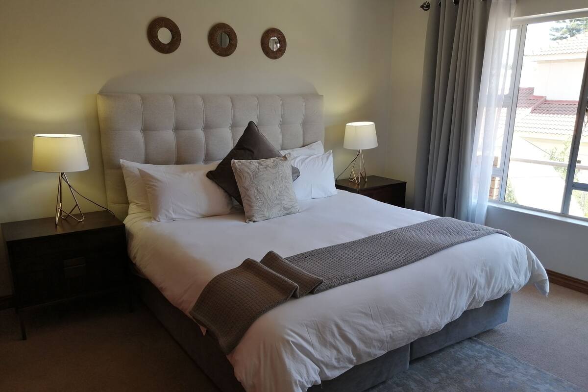Una cama bien tendida con un cabecero capitonado, dos mesitas de noche con lámparas, y tres espejos decorativos colgados en la pared.