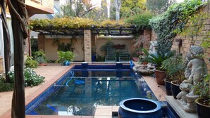 Una piscina rectangular con borde azul, rodeada de plantas en macetas y una estatua de piedra.