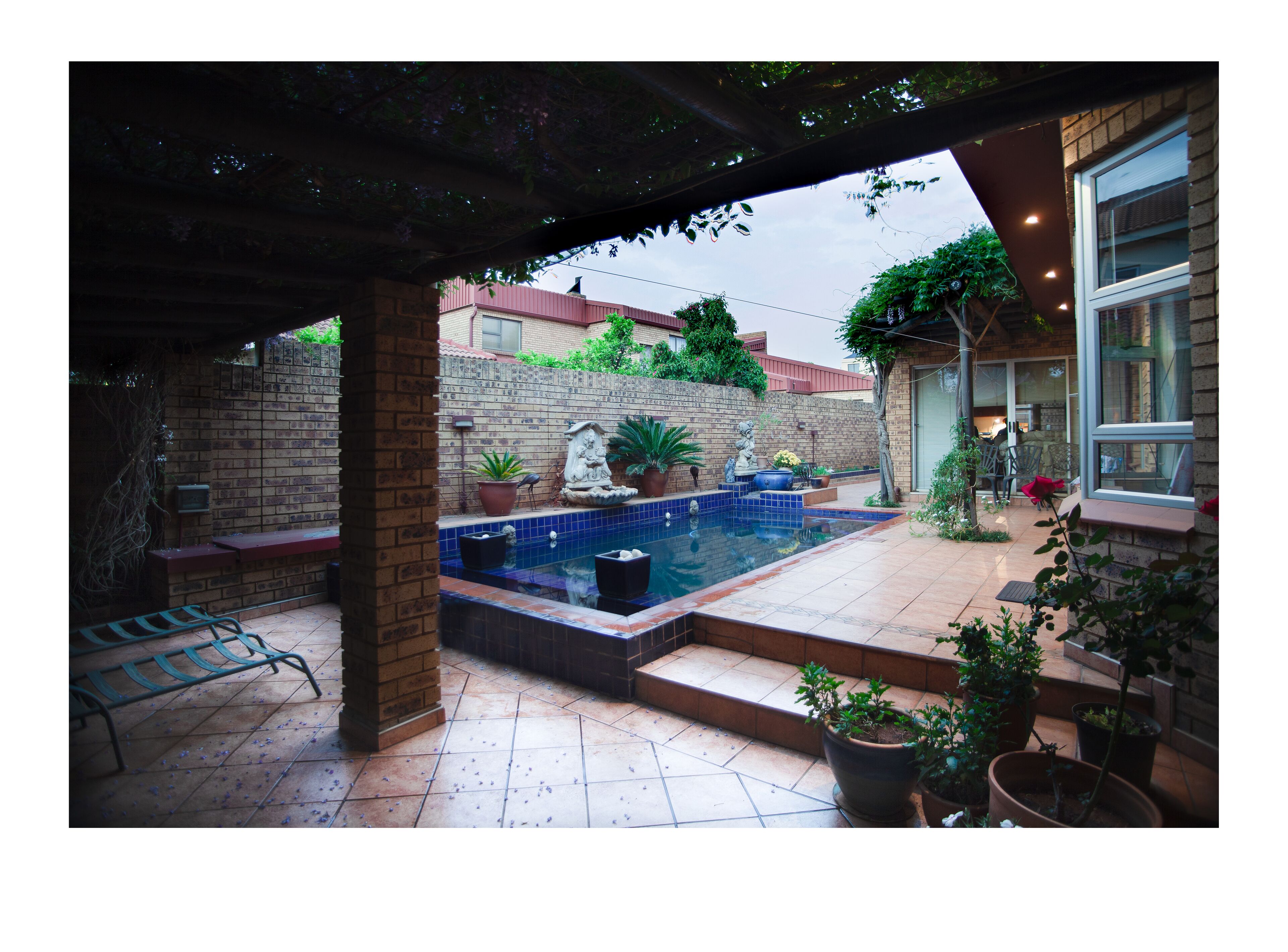 Un espacio exterior techado con piscina, plantas en macetas y un muro de ladrillo.