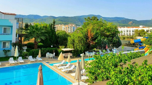 A pool area with lounge chairs and umbrellas, surrounded by greenery and buildings in the background. - Melodi Club Hotel (Kusadasi)