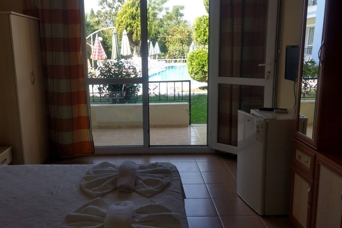 A hotel room with a bed, a small table, a refrigerator, and a sliding glass door leading to an outdoor area with a pool and umbrellas.