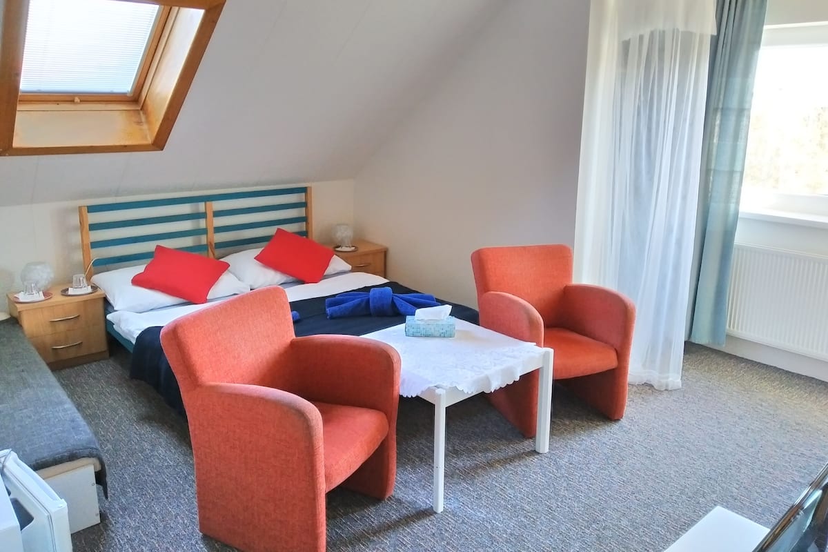 A bedroom with a bed, two orange chairs, a small table, a nightstand, and a window with curtains.