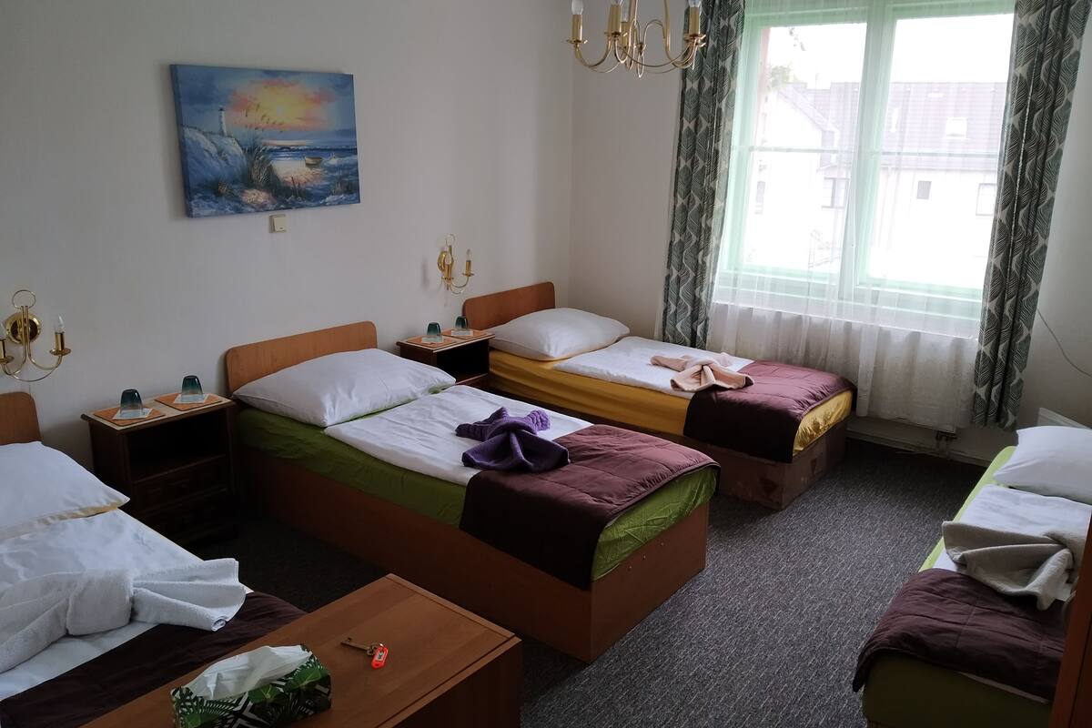 A hotel room with two single beds, a wooden nightstand with a lamp, a painting on the wall, and a window with patterned curtains.